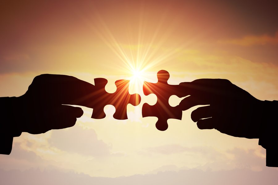 Zwei Hände fügen vor einem Sonnenuntergang zwei Puzzleteile zusammen.